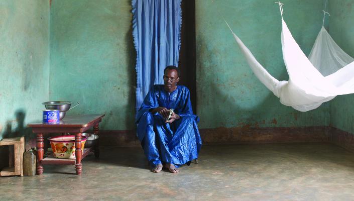 Ogobaro Doumbo sits on a low chair in his home. A mosquito net hangs to his left.