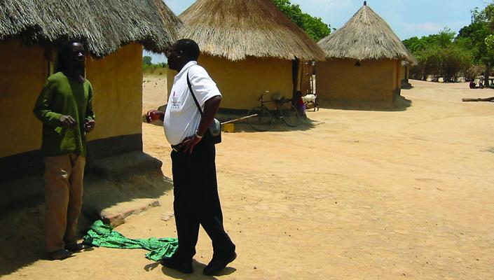 a CREATE worker talks with a community member outside a mud and thatch house