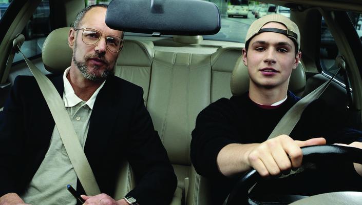 a teen wearing a backward baseball cap sits behind the steering wheel; his father sits in the passenger seat.