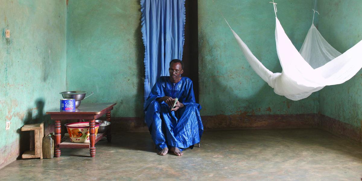 Ogobaro Doumbo sits on a low chair in his home. A mosquito net hangs to his left.