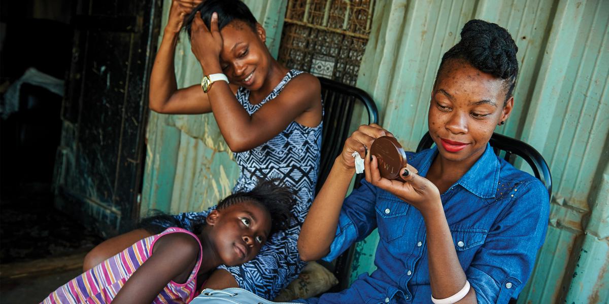 two young women, one fixing her makeup and the other fixing her hair, smile as a younger girl looks on