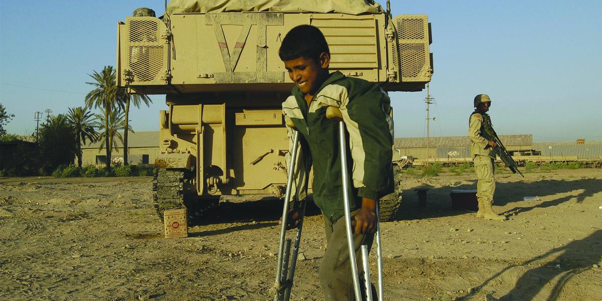 A young teen on crutches walks by a military vehicle guarded by a soldier