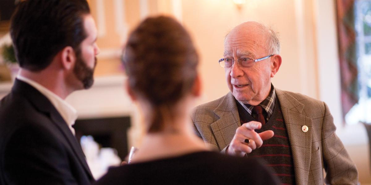 Clive Shiff, wearing a shirt, tie, and jacket speaks with a man and woman whose backs are to the camera