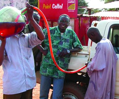 3 men in Chad fill their white pickup's gas tank by siphoning fuel from a glass bottle