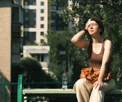 A woman sits on a city park bench while wiping her brow in the midday heat.