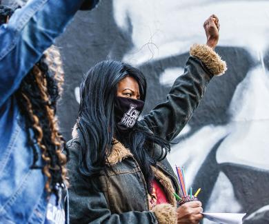 Black women standing in front of a George Floyd mural with raised fists.